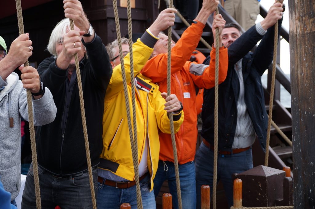 Fünf Männer ziehen an Seilen um Segel zu setzen